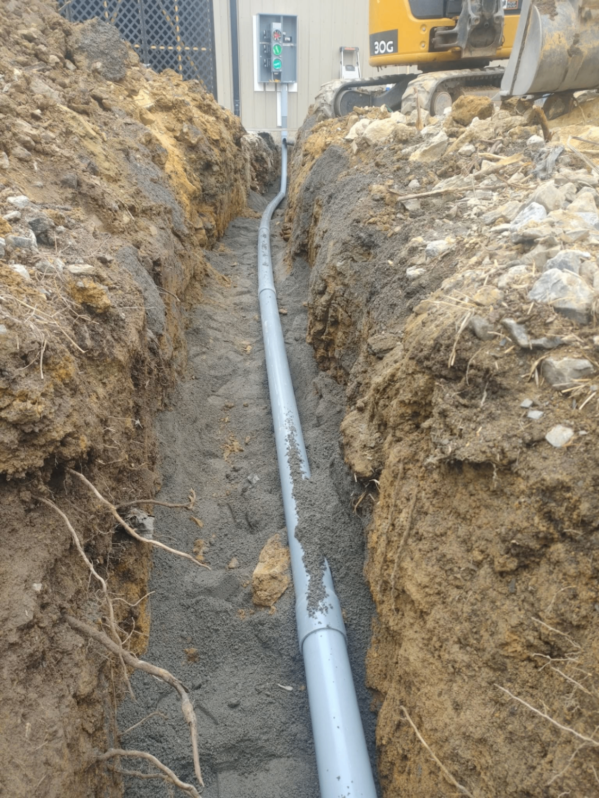 Underground PVC conduit run in an open trench beside a commercial building with excavator