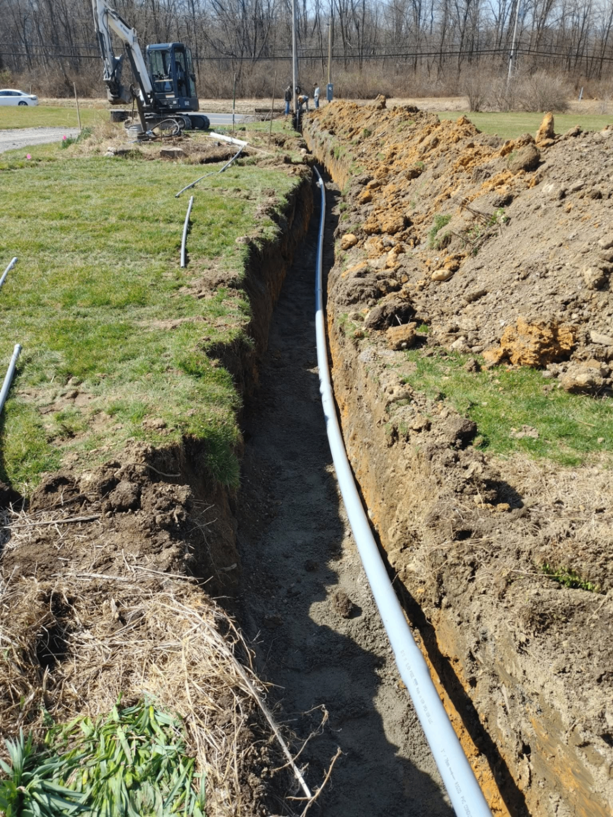 Long open trench with underground PVC conduit run and excavator parked roadside