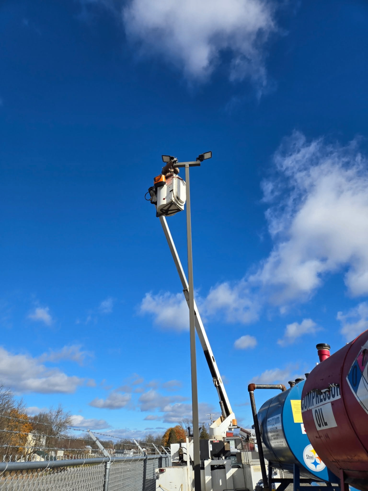 Technician in a bucket lift installing parking lot light fixtures on top of a tall pole under a blue sky