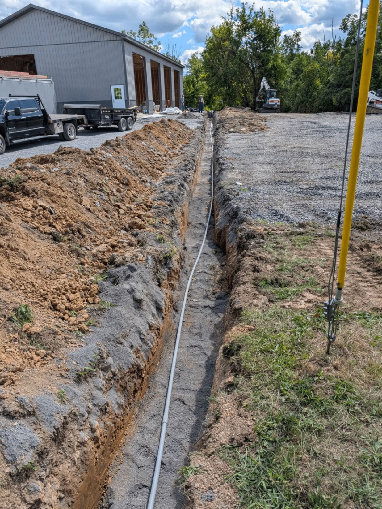 Long open trench with underground conduit running alongside a commercial pole building and excavator